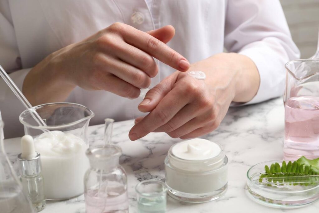 Person trägt Creme aus einem Tiegel auf den Handrücken auf, umgeben von Hautpflege-Produkten und Laborgefäßen